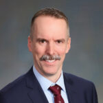 Bob Curran Headshot_web Headshot of a person with short hair and a mustache, dressed in a suit and tie, against a plain background.